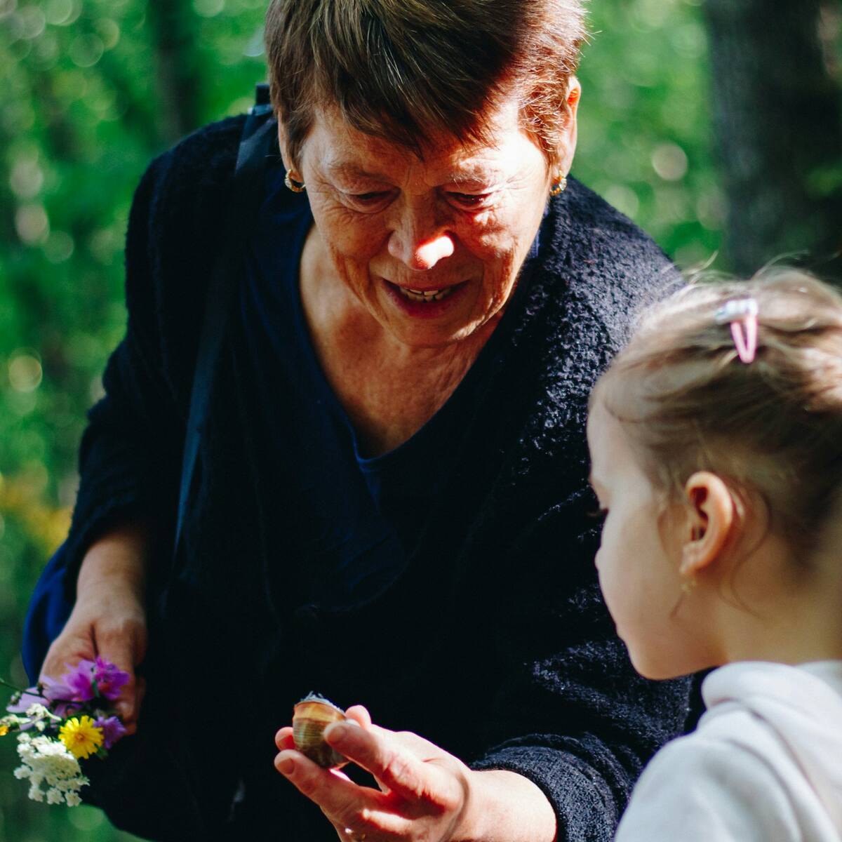 A grandmother shows her granddaughter a snail shell on a walk in the woods